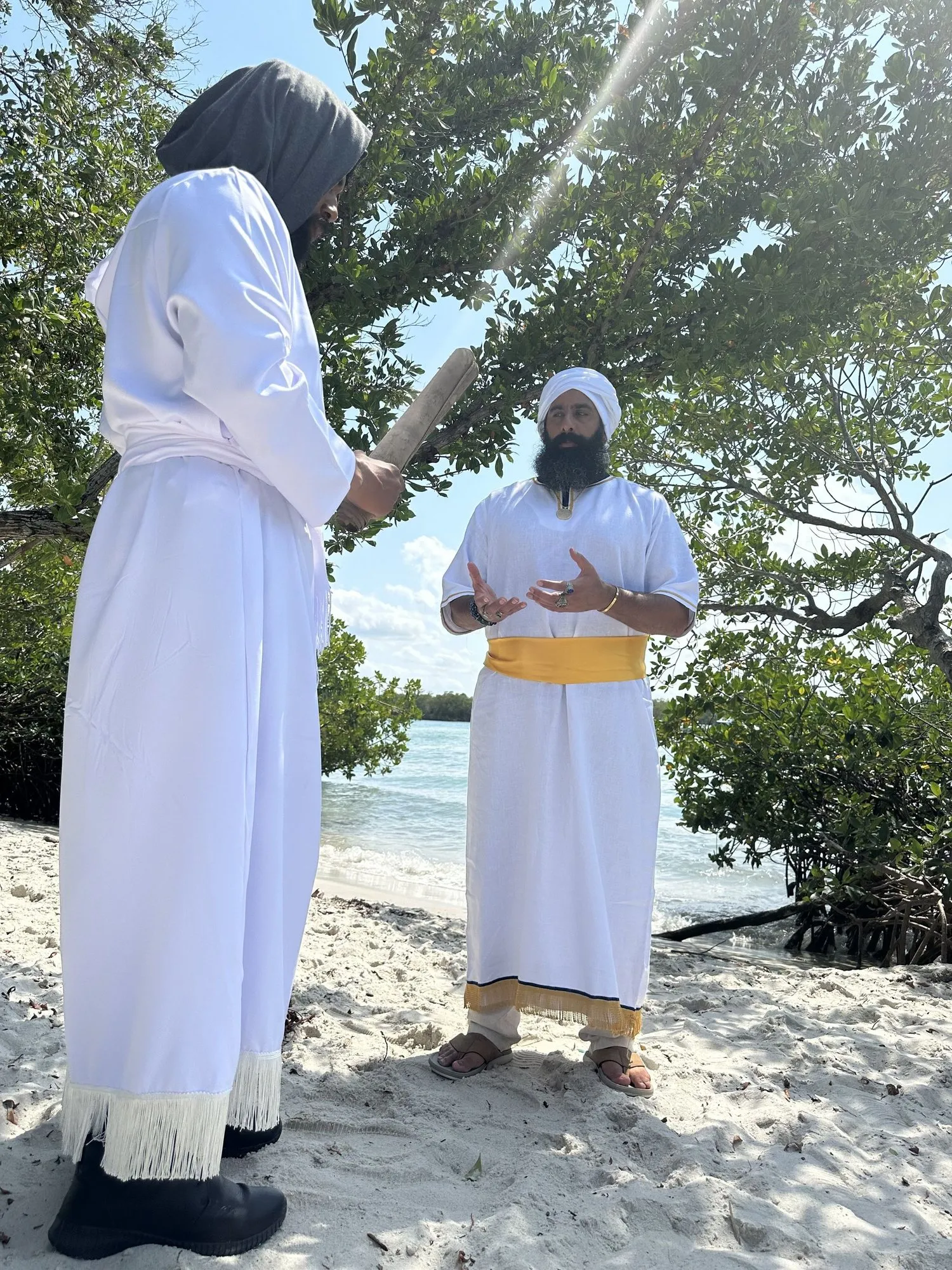 Two men in white robes on a beach, one lifting his hand over the other in blessing.