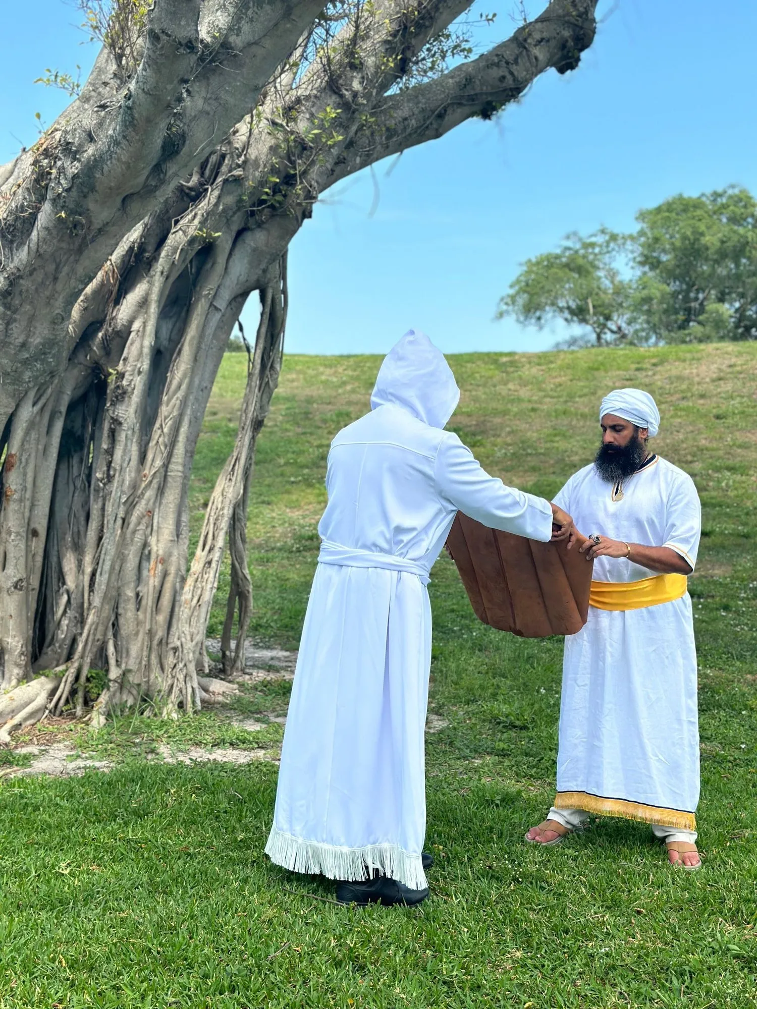 Two men in white priestly garments meet beneath a great tree, one presenting a gift to the other.