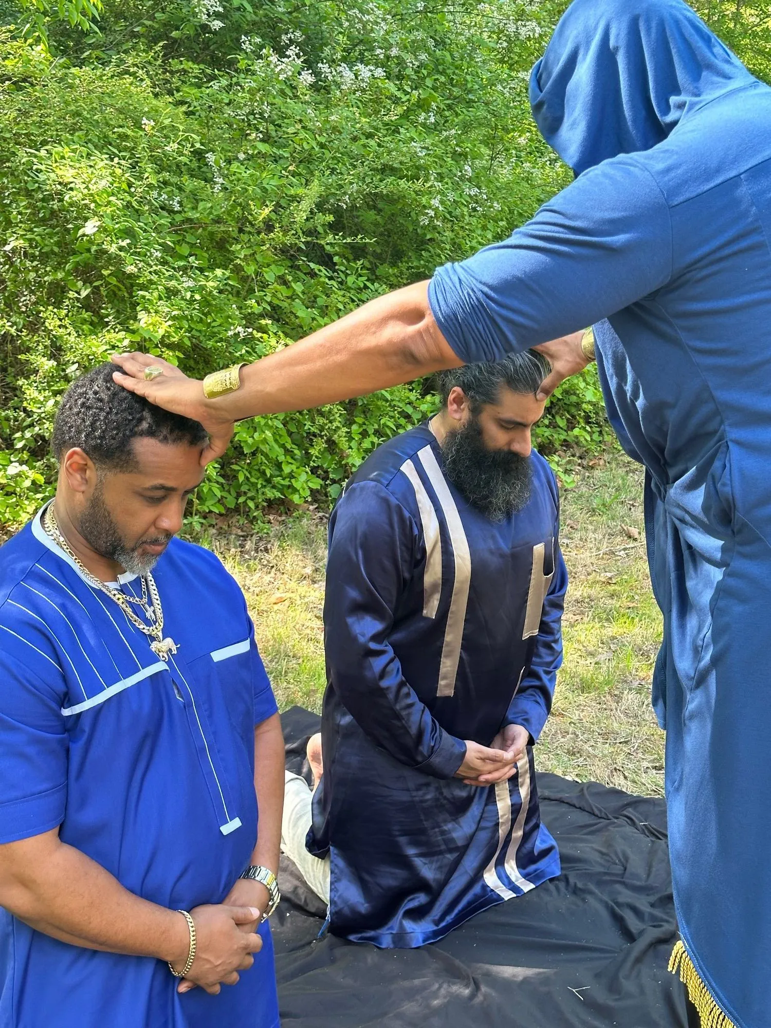 Two men kneel on the grass as hands are laid on their heads in blessing.
